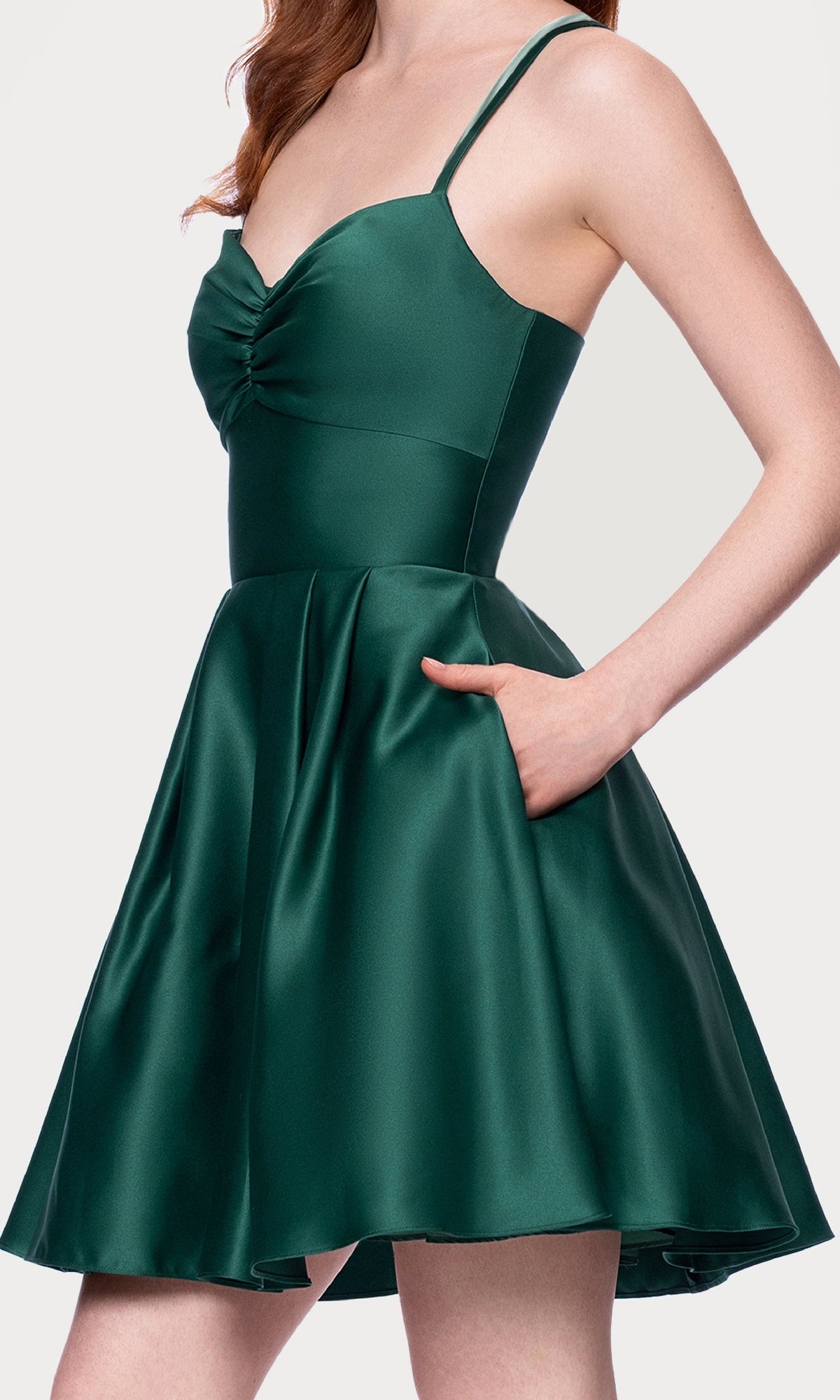 A woman poses with one hand in the pocket of the Hunter Green Mini Hoco Dress: Blondie Nites 4927BN, a sleeveless satin dress featuring a lace-up back, spaghetti straps, and flared skirt.